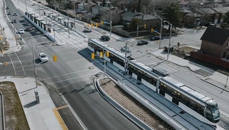 The image shows a modern urban intersection featuring a dedicated tramway line. At the centre, a sleek articulated tram in white and black runs on segregated tracks bordered by concrete curbs. The tram, composed of multiple cars, is equipped with pantographs for overhead electric power supply. On the left, there is a station with raised platforms, sheltered by metallic structures and fitted with information panels. Several traffic lights and road signs regulate the movement of vehicles and pedestrians.