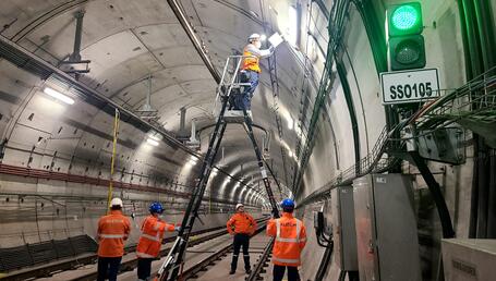 The image shows a team of technicians working inside a large railway tunnel. The perfectly curved concrete walls stretch out into the distance and are lit by a series of lightings attached to the ceiling. In the centre, a technician wearing an orange vest and safety helmet stands high up on a large articulated ladder to work on a box or electrical component located above.